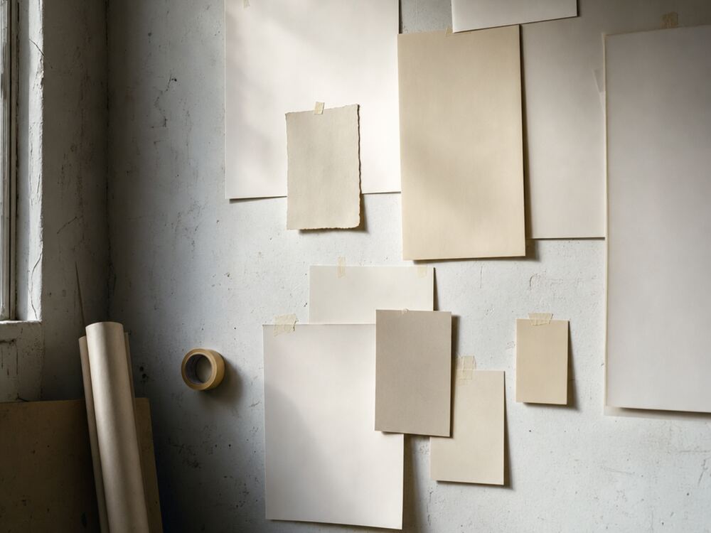 Pliegos de papel en blanco sujetos con cinta sobre la pared de un taller, bajo luz de ventana.